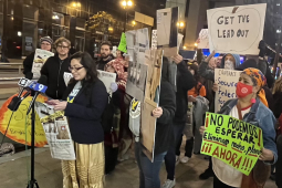 a speaker at a mic set up on the sidewalk is backed up by people holding signs about lead in schools