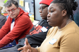 A Black woman in a tan shirt speaks while three men, white and Black, look on.