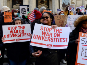 A big crowd, bundled against cold, stands in front of a building with columns. Foregrounded are four women, one with beaming smile, one looking up warmly at her. All hold PSC-CUNY signs that say variously, "Stop the War on Universities," "Save Research," and "Science is for the People." Other handmade signs, a UAW logo, and an upside-down American flag are visible in the crowd behind htem.