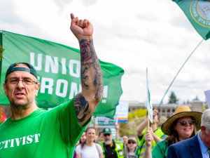 A man in a backwards green ballcap and an AFSCME green T-shirt printed with the word "Activist" raises his heavily tattooed arm up to the sky. Behind him other marchers are visible, crossing the capitol campus, some with signs (not readable) and one waving a Washington state flag on a tall pole.