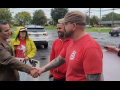 La captura de pantalla muestra a Israel Cervantes, a la izquierda, con una chaqueta de cuero marrón, estrechando la mano de Sean Crawford, a la derecha, con una camiseta roja de la UAW y una gorra de béisbol, mientras otros activistas con carteles hechos a mano observan con aprobación. Están de pie en un estacionamiento después de llover.
