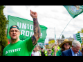 A man in a backwards green ballcap and an AFSCME green T-shirt printed with the word "Activist" raises his heavily tattooed arm up to the sky. Behind him other marchers are visible, crossing the capitol campus, some with signs (not readable) and one waving a Washington state flag on a tall pole.