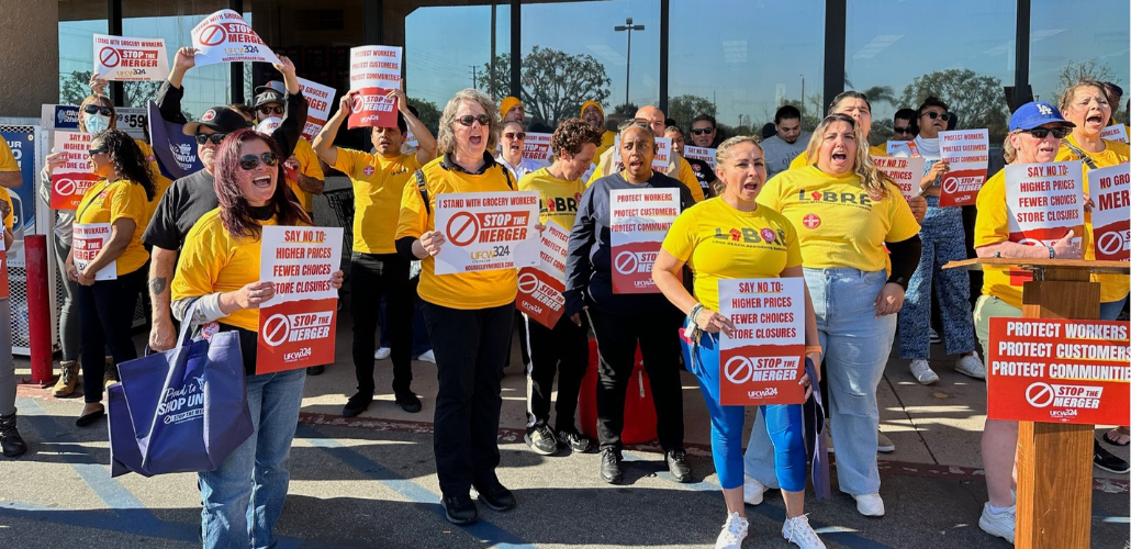 A crowd of people, mostly women, racially varied, in yellow T-shirts (some say UFCW, some say LIBRE) stand outside a grocery store, holding printed red-and-white signs, shouting together. There is a podium. Printed signs have UFCW Local 324 logo and such messages as "Stop the merger," "Say no to higher prices, fewer choices, store closures," and "Protect workers, protect customers, protect community."