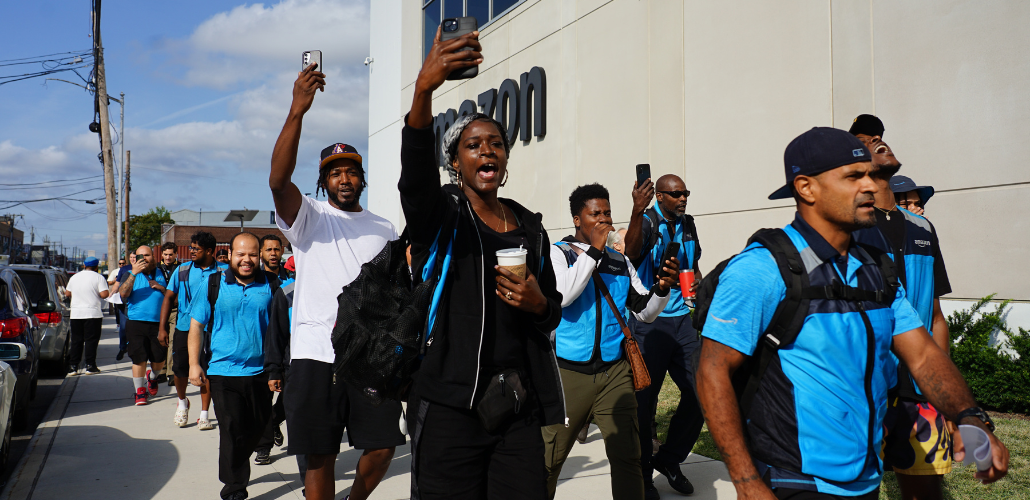 Workers, some in blue vests, march outside along an Amazon building. Some are holding their phones aloft, recording. Many are Black. They look like they are striding confidently and chanting.