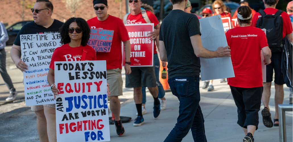 Strikers picket in red shirts. Most prominent is a woman with a sign: "Today's lesson: equity and justice are worth fighting for." Behind her is a man with a partially obscured sign listing "Part-time jobs I've worked so I can afford to be a teacher in Massachusetts" including Lyft driver, nightclub bouncer, Doordash, adjunct.
