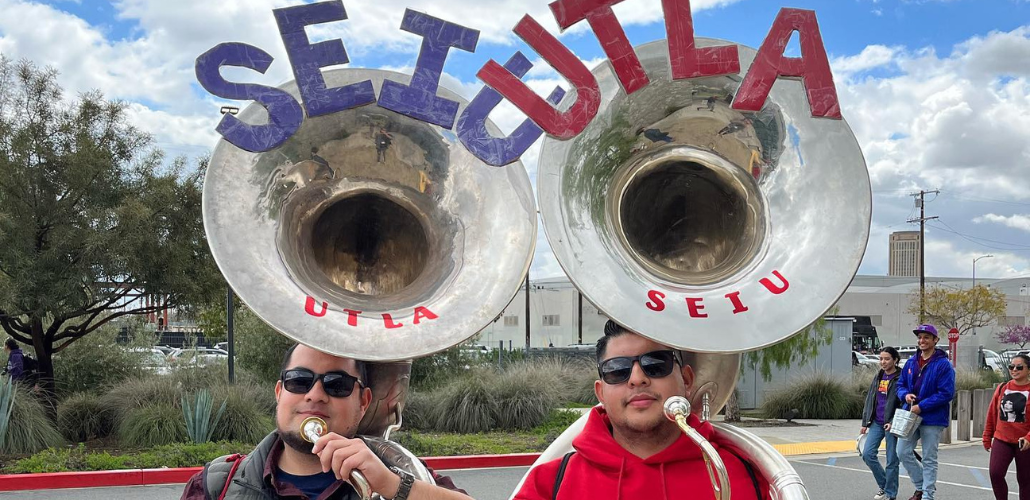 Two men in matching expressions and sunglasses stand elbow to elbow, facing the camera, holding souzaphones. One instrument has "SEIU" in purple cutout letters at the top of the horn, and "UTLA" in smaller red sticker letters below the opening; the other has the reverse. One man wears a purple shirt, the other a red hoodie. Behind them is a sunny sky and several other picketers approaching.