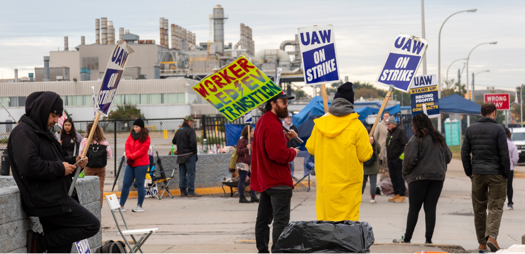 Workers in heavy coats stand around picketing in front of a big gray factory. One neon-colored handmade sign says "Worker-led EV transition" with a lightning bolt. Other signs are printed and say "UAW on strike" or "UAW, Stand up, Record Profits, Record Contracts"