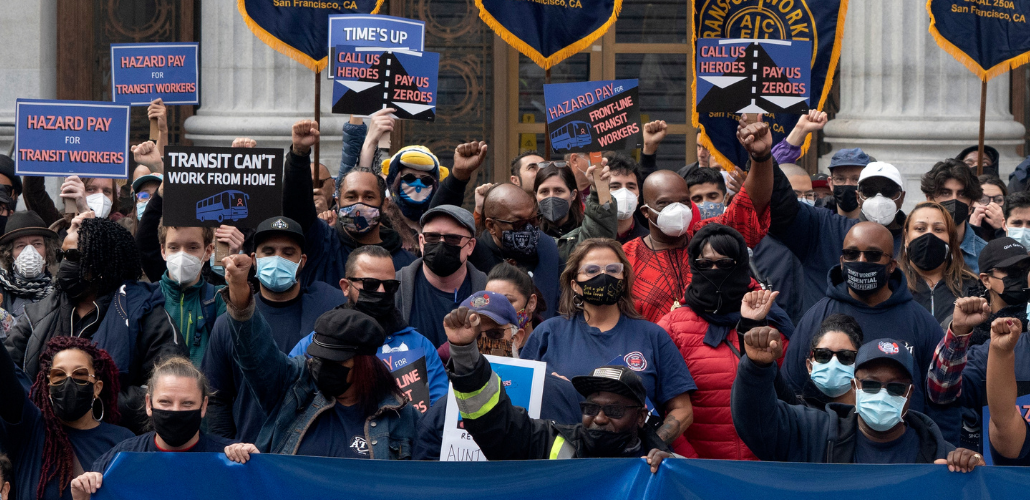 A diverse crowd, all masked, some with fists in the air, outdoors on steps. Printed signs read: "Transit can't work from home," "Hazard pay for transit workers," and "Call us heroes, pay us zeroes"