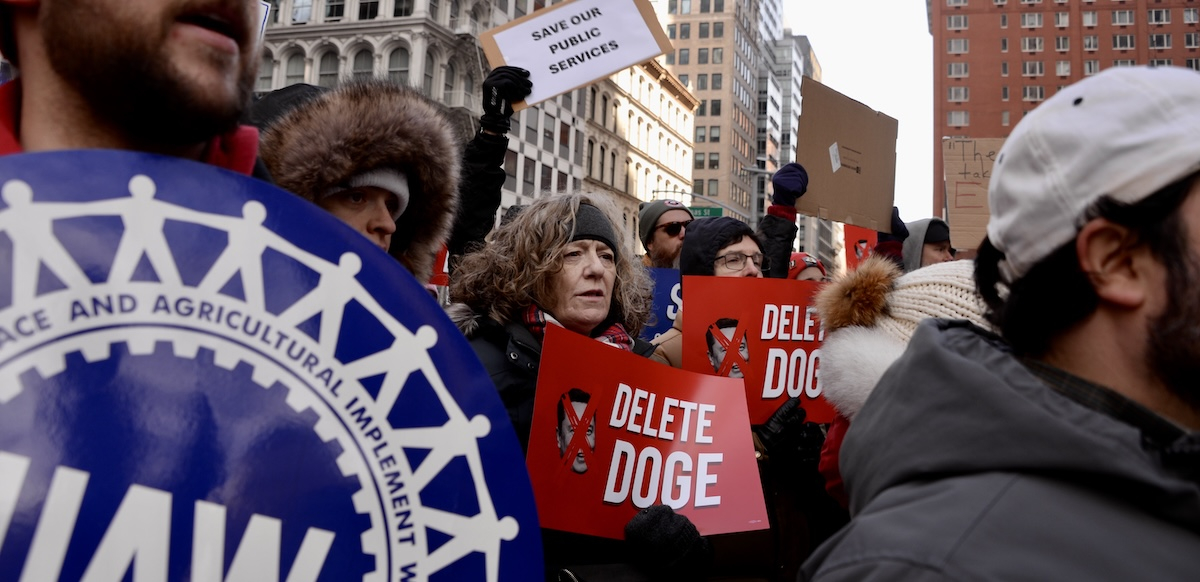 A crowd of people hold red and white signs saying “Delete Doge”