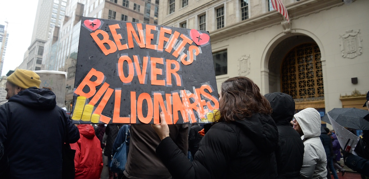 Marchers walk away from the camera with a giant orange on black sign, “Benefits over Billionaires”