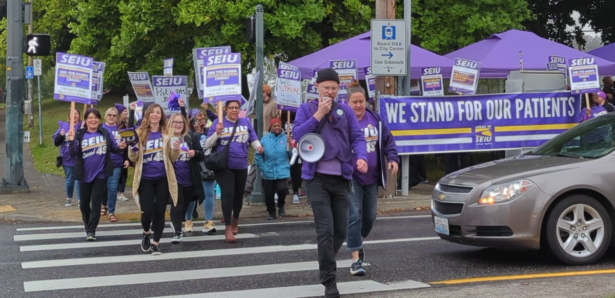 Workers in purple SEIU Local 49 T-shirts march across a street. Some carry picket signs that say SEIU and some other words too small to read. A large purple banner says "We stand for our patients." The person in front is talking into a bullhorn and others appear to be chanting.