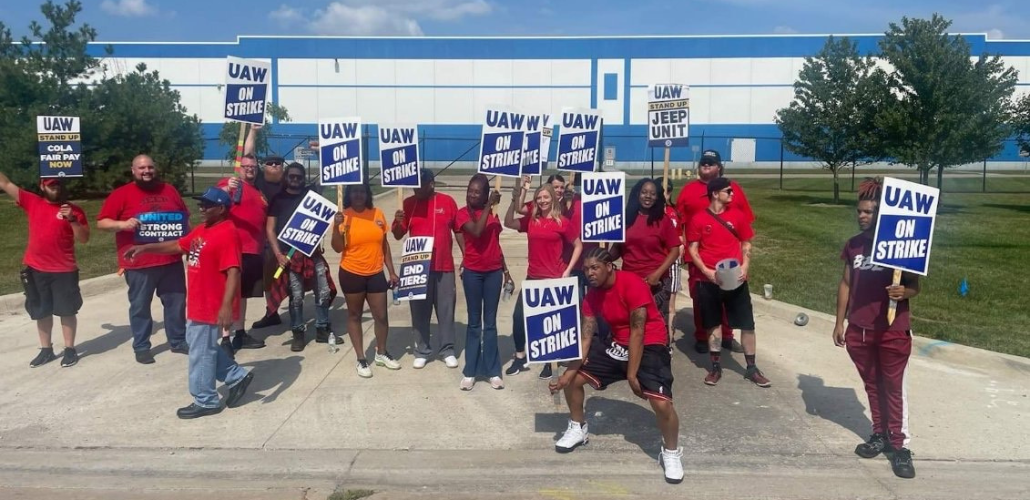 A group of Black and white men and women, most in red T-shirts, stand in front of the plant gates. Many carry blue and white "UAW on strike" picket signs.