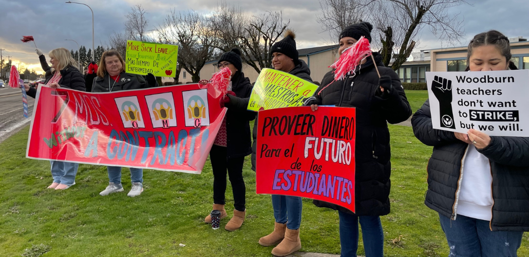 People stand on grass with signs. A red banner says "Seven months without a contract, OMG!" with stylized images of Edward Munch's painting "The Scream." Another sign says "More sick leave" in English, Spanish, and Russian. Another says, in Spanish,"Provide money for the future of students." Another says "Woodburn teachers don't want to strike, but we will."