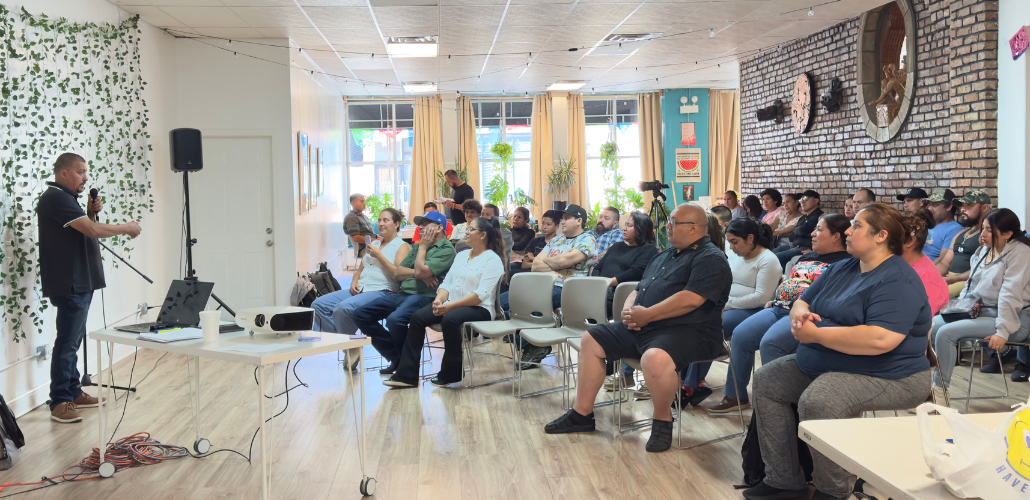 In a bright sunny room, a man in a black polo shirt and jeans stands on the left, speaking into mic, while about 30 people seated in chairs on the right listen. Most or all appear Latino/a.