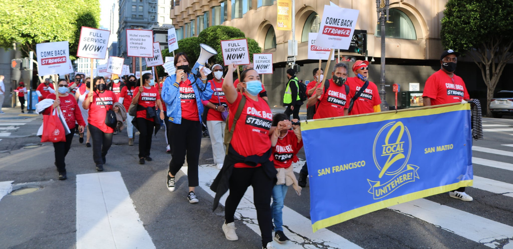 Workers march in downtown streets behind a big blue banner that says "UNITE HERE Local 2." They are wearing red T-shirts that say "Come back stronger" and some carry picket signs saying "Full service now!" or "Hotel rooms should be cleaned every day." Most wear masks, one holds a bullhorn, and there is a child near the front of the crowd.