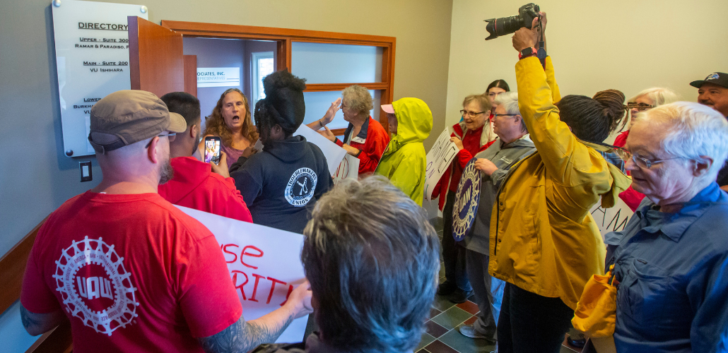 At least a dozen activists crowd up to the door of a VU Manufacturing office, where a company flack is gesuring widely and trying to fend them off. One activist is holding a camera aloft, others holding signs or wearing UAW or Labor Notes gear. A woman
