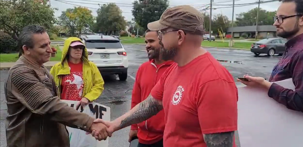 Screenshot shows Israel Cervantes on the left, in a weathered brown leather jacket, shaking hands with Sean Crawford, right, in a red UAW T-shirt and a baseball cap, while a few other activists carrying handmade signs look on approvingly. They're standing in a parking lot after a rain.
