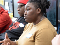 A Black woman in a tan shirt speaks while three men, white and Black, look on.
